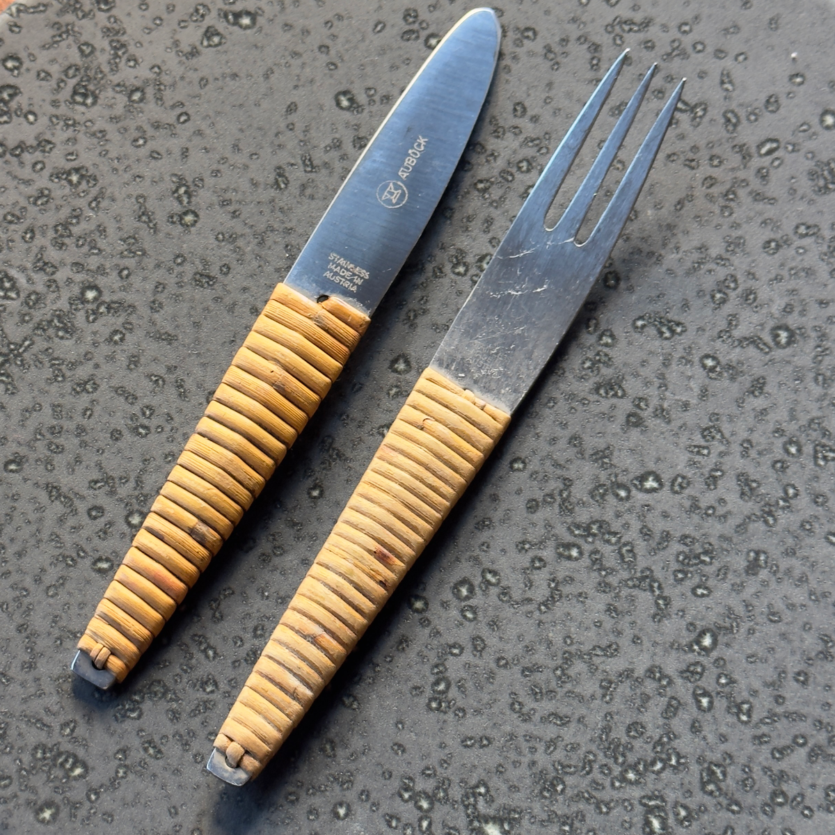 A vintage 1950s cutlery set by Carl Auböck, featuring a knife and fork with sleek steel blades and distinctive cane-wrapped handles. This mid-century design blends functionality with artisanal craftsmanship, showcased at Sigmar London.