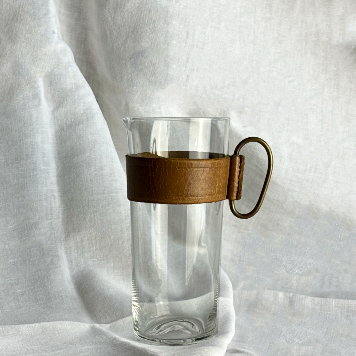 Vintage glass pitcher by Carl Auböck II, featuring a leather band and brass handle. This Austrian design exemplifies Auböck’s signature blend of functional craftsmanship and modernist elegance, available at Sigmar.