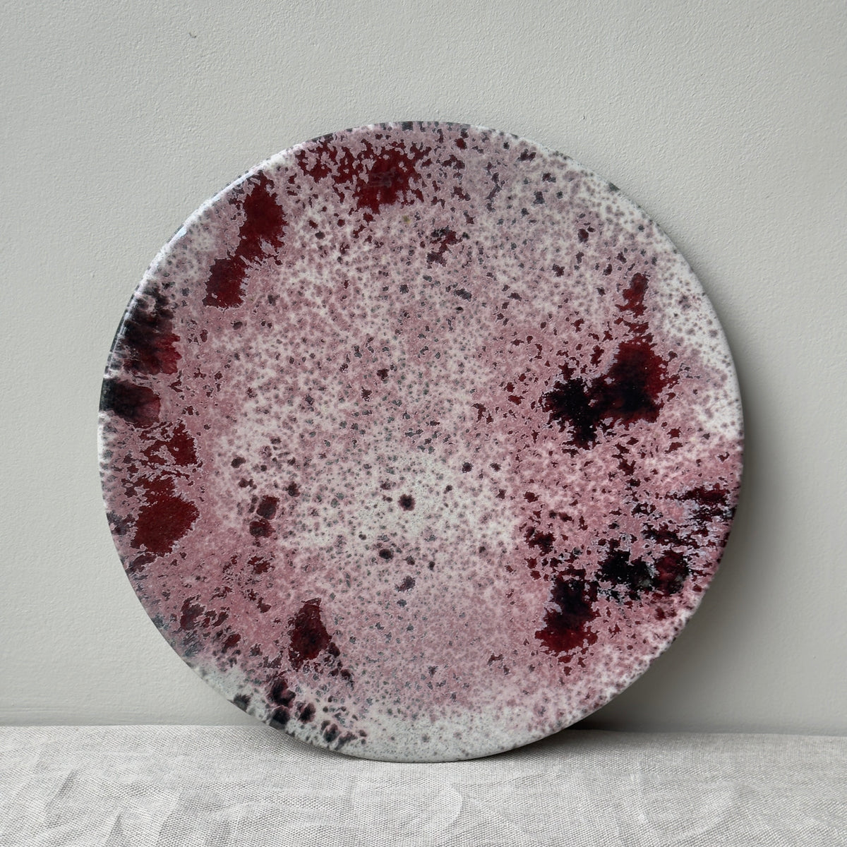 A red stoneware art piece platter made and designed in Denmark shown against a grey wall on a flat linen surface. 